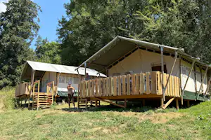 Großes Zelt auf Holzpodest mit Geländer, daneben eine Person vor dem Eingang, umgeben von Bäumen und Wiese.