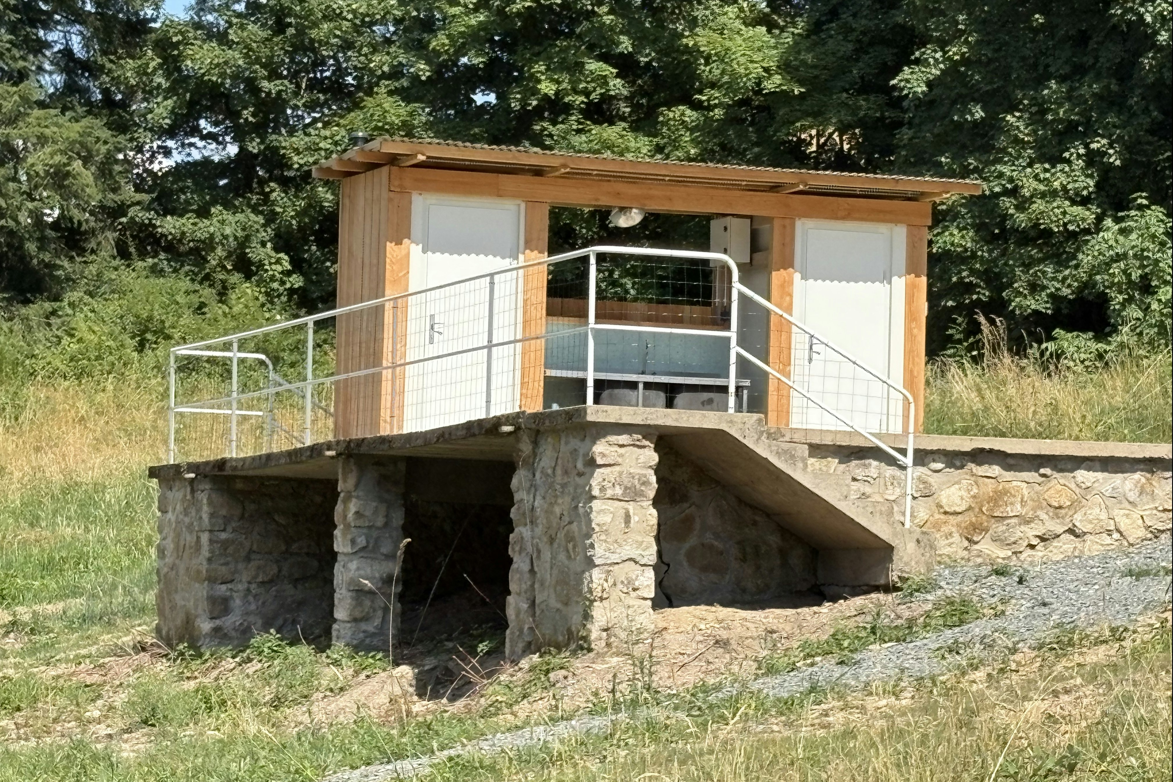 Holzüberdachter Sanitärbereich mit Metallgeländer auf einem steinernen Fundament, umgeben von Wiese und Bäumen.