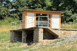 Holzüberdachter Sanitärbereich mit Metallgeländer auf einem steinernen Fundament, umgeben von Wiese und Bäumen.