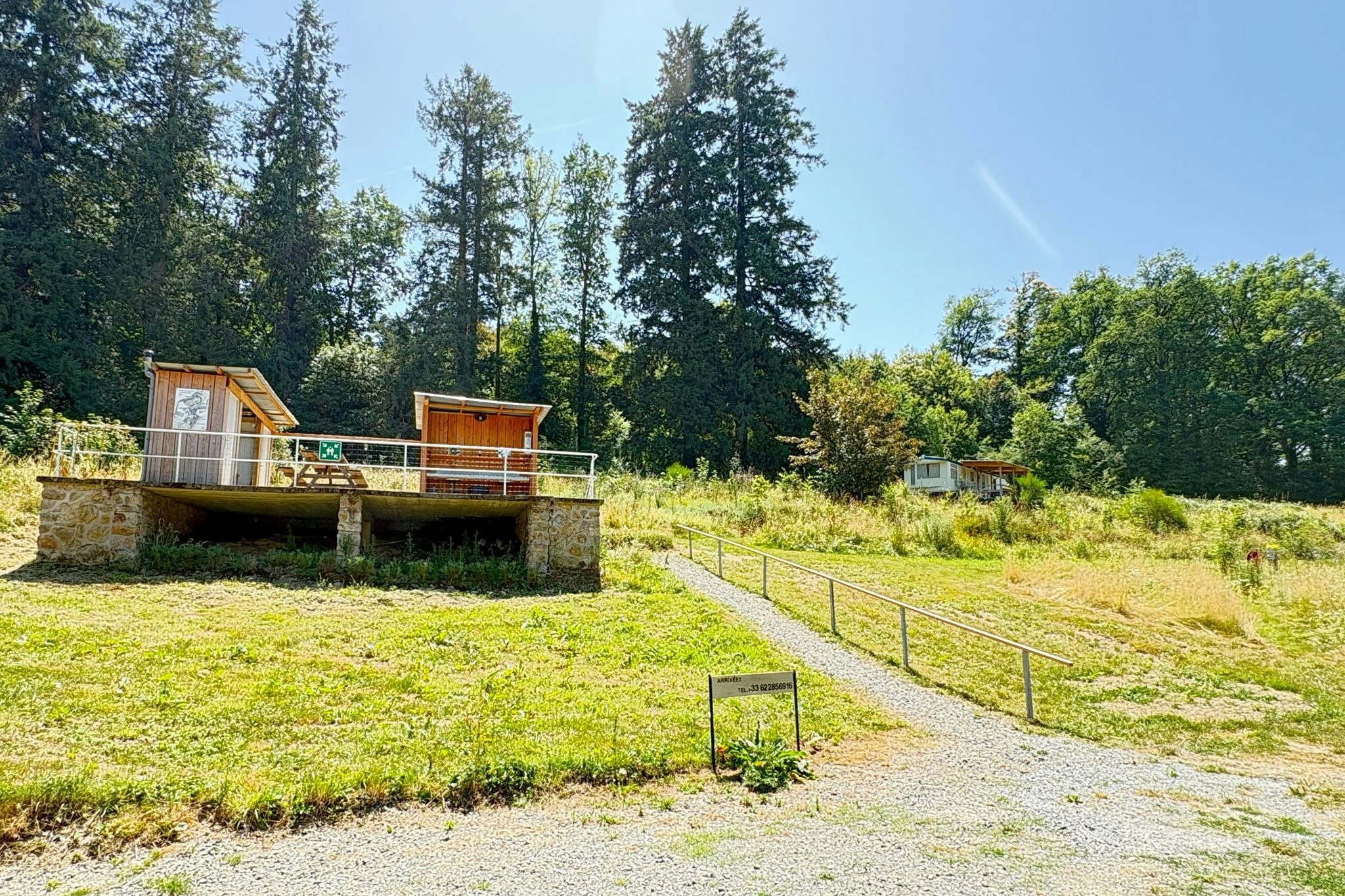 Holzhütten auf einem Campingplatz mit Terrasse, umgeben von Wiesen und hohen Bäumen, daneben ein Kiesweg.
