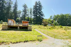 Holzhütten auf einem Campingplatz mit Terrasse, umgeben von Wiesen und hohen Bäumen, daneben ein Kiesweg.