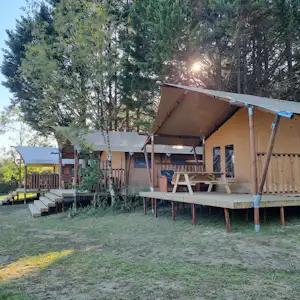 Zelt mit Holzterrasse und Sitzbank auf einem Campingplatz, umgeben von Bäumen und Rasenfläche.