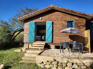Holzbungalow mit blauen Fensterläden, Terrasse mit Tisch und Stühlen sowie Sonnenschirm, umgeben von Garten und Büschen.