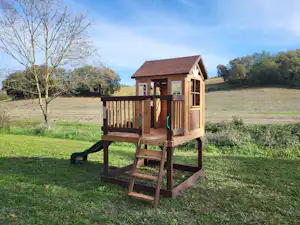 Spielhaus für Kinder auf einem Campingplatz, mit Holztreppe, Veranda und Blick auf Wiese und Bäume.