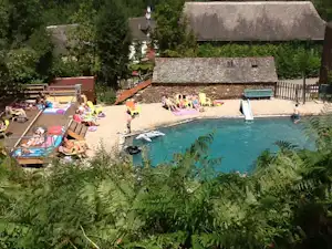 Schwimmbecken mit türkisfarbenem Wasser, Liegen und Sonnenschirmen auf dem Kiesplatz, umgeben von Bäumen und Gebäuden.