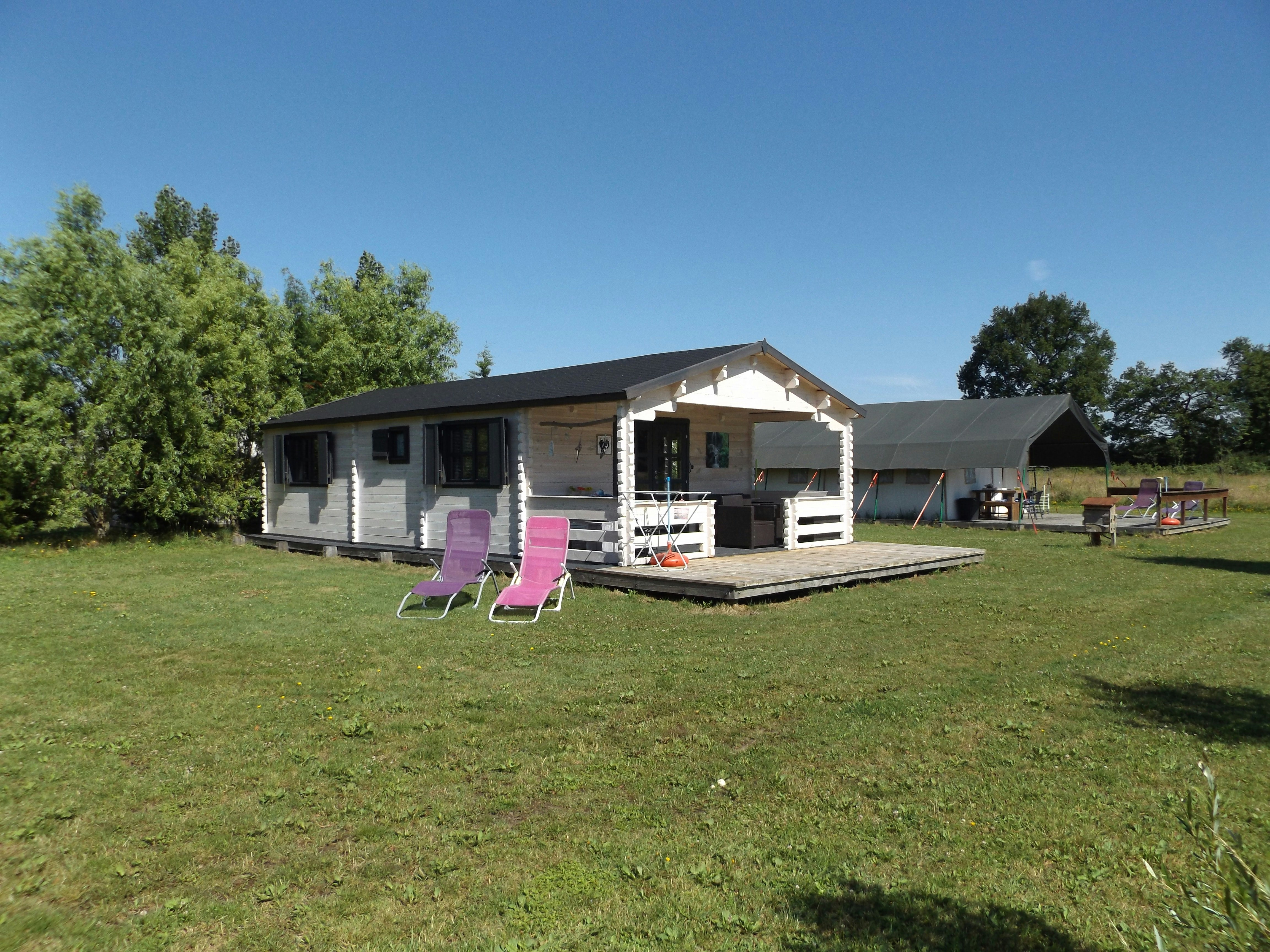 Wohnwagen mit Terrasse, zwei Gartenstühlen und Tisch auf einem gepflegten Rasen, im Hintergrund Bäume und ein weiteres Zelt.