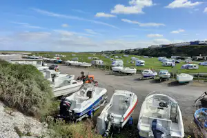 Reihe von Wohnmobilen und Booten auf Stellplätzen am Meer, mit Blick auf Küste, Grasflächen und Wohnwagen im Hintergrund.