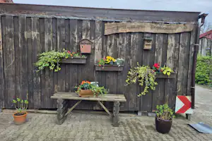 Holzfassade mit Bank und Blumenkästen, mehrere Töpfe mit Pflanzen vor einer Campinghütte, daneben ein Schild mit rotem Pfeil.