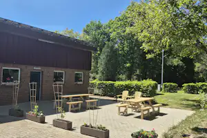 Terrasse am Campingplatz mit Holztischen und Stühlen vor einem Backsteingebäude, umgeben von Hecken und Bäumen.