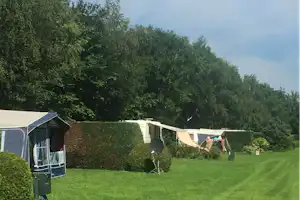 Zelt und Wohnwagen auf einem grünen Campingplatz, Hecken und Bäume im Hintergrund, Wiese mit Campingstühlen.