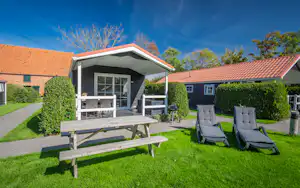 Ferienhaus mit Terrasse, Markise und großen Fenstern, davor Holztisch, Liegestühle und Grill im gepflegten Garten.