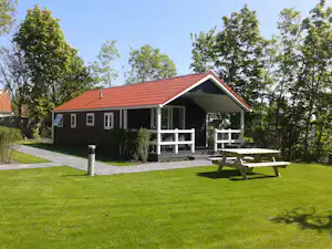 Ferienhaus mit rotem Ziegeldach, Terrasse und Gartenmöbeln vor grünen Bäumen, mit gepflastertem Weg und Rasen.