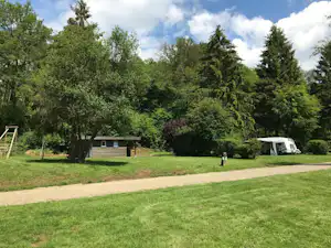 Zelt auf einem Campingplatz im Grünen, mit Kiesweg, Bäumen und einem kleinen Holzgebäude im Hintergrund.