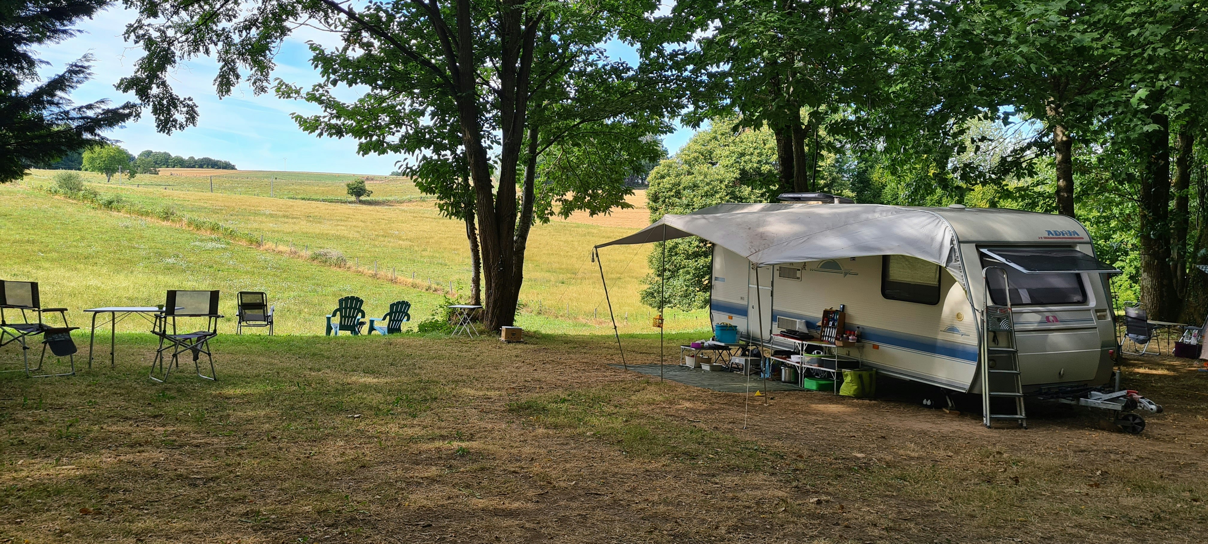 Wohnwagen auf einem Campingplatz im Grünen, mit Vorzelt, Campingstühlen und Tisch unter Bäumen am Rand einer Wiese.