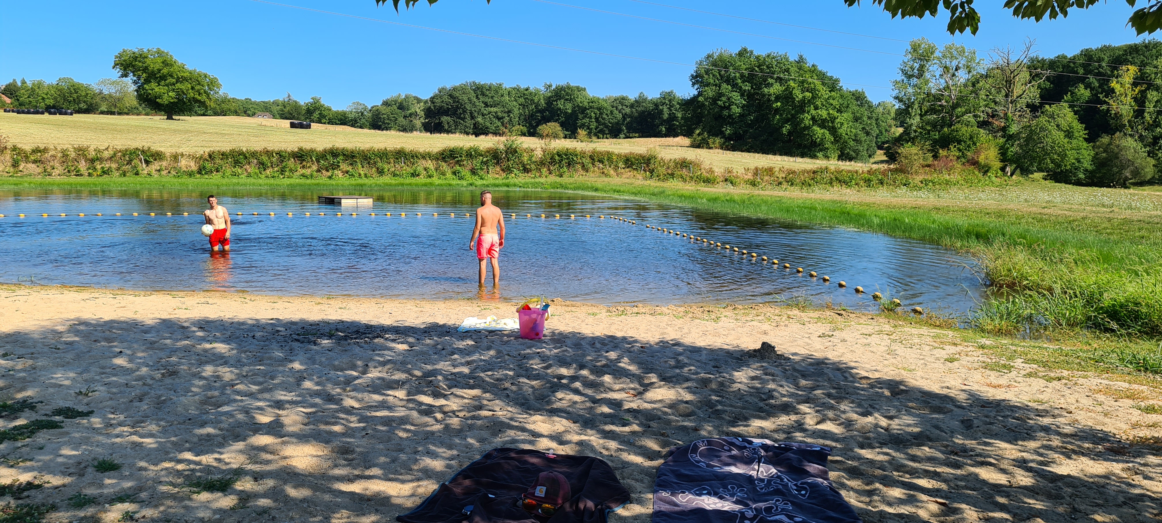 Badestrand am See mit zwei Personen im Wasser, flachem Ufer und Bäumen am Rand.