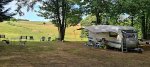 Wohnwagen mit Vorzelt auf einem Campingplatz, Sitzgruppe und Tisch unter Bäumen, Wiese und Zaun im Hintergrund.