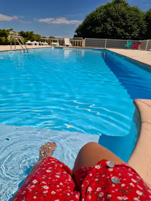Blick auf das blaue Schwimmbecken mit Leiter, im Hintergrund liegen weiße Liegestühle und bunte Stühle am Zaun.
