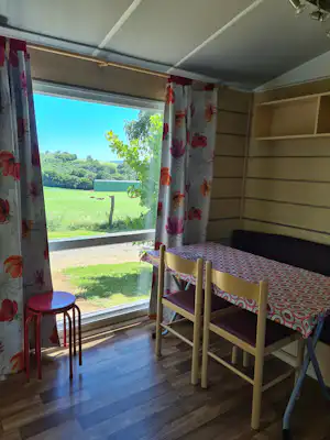 Innenansicht eines Wohnwagens mit Esstisch und Stühlen am Fenster, Blick auf Wiese und Bäume.
