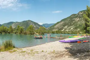 Blick auf den Campingsee mit Kajaks am Ufer, Schwimmern im Wasser und Bergen im Hintergrund.