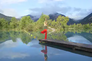 Person macht Yoga auf einem Holzsteg am See, mit Spiegelung im Wasser und Bergen im Hintergrund.