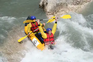 Zwei Personen im gelben Schlauchboot mit Paddeln und blauen Helmen auf schäumendem Fluss, Felsen im Hintergrund.