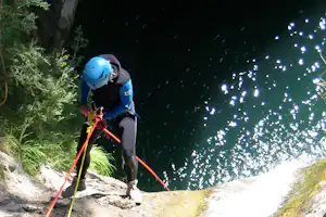 Kletterer mit blauem Helm und Klettergurt am Fels, Seile und Karabiner, Wasser spritzt im Hintergrund.