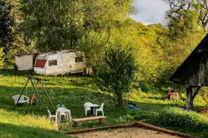 Campingwiese mit Zelt, Picknicktisch und Stühlen, umgeben von Bäumen, im Hintergrund Berge und Nebel.