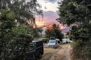 Campingweg zwischen Wohnmobilen und einem kleinen Wohnwagen, umgeben von Bäumen, im Abendlicht.
