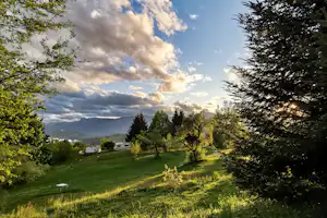 Grüne Campingwiese mit Bäumen, kleinen Wohnwagen und Blick ins Tal unter wolkigem Himmel.