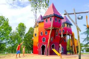 Buntes Spielhaus aus Holz mit Türmen und Fenstern im Freien, daneben ein Spielgerät und Bäume im Hintergrund.