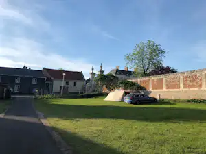 Campingplatz mit Rasenfläche, gepflastertem Weg und mehreren Gebäuden, im Hintergrund ein Auto neben einer Mauer.