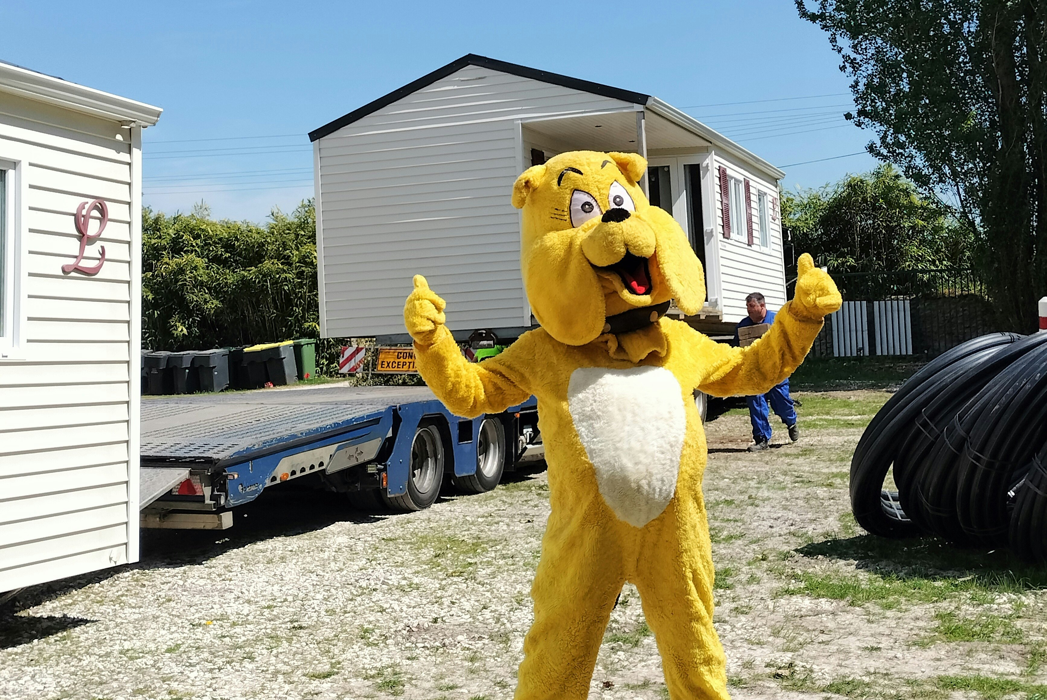 Gelbes Maskottchen mit offenen Armen vor einem Wohnwagen-Anhänger neben einem weißen Gebäude und Hecken.