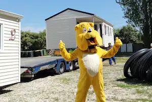 Gelbes Maskottchen mit offenen Armen vor einem Wohnwagen-Anhänger neben einem weißen Gebäude und Hecken.