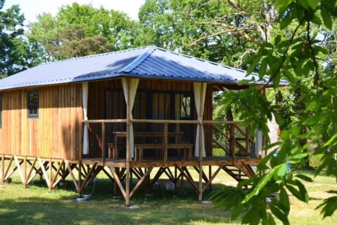 Holzbungalow mit Veranda, Vorhängen und Geländer auf Stelzen, umgeben von Bäumen und Rasen im Campingbereich.