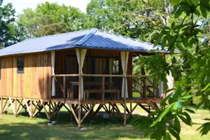 Holzbungalow mit Veranda, Vorhängen und Geländer auf Stelzen, umgeben von Bäumen und Rasen im Campingbereich.