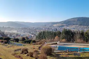 Blick über einen Campingplatz mit Wiese, Stellplätzen und einem blauen Pool, umgeben von Bäumen und Hügeln im Tal.