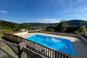 Großes Camping-Schwimmbecken mit Leiter, umgeben von Terrasse und Zäunen, im Hintergrund Hügel und Häuser im Tal.
