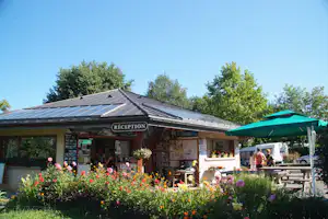 Rezeption am Campingplatz mit Schild „RÉCEPTION“, Blumenbeet und grünen Sonnenschirmen vor dem Gebäude.