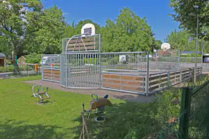 Kinderbett mit Holzlatten und Metallgeländer auf dem Campingplatz, im Hintergrund Bäume und weitere Unterkünfte.