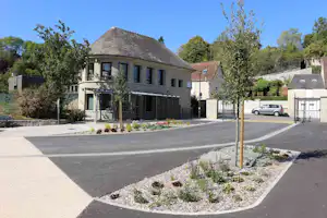 Campinghaus mit Terrasse und Treppe, gepflasterter Einfahrt und Parkplatz mit Auto, Hecken und Bäumen im Hintergrund.