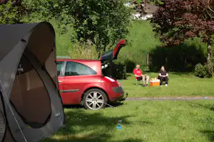 Zelt und rotes Auto auf einem grünen Campingplatz, daneben Rasenweg und zwei Personen mit Getränkekisten.