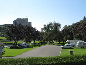 Campingplatz mit Wohnwagen und Auto am Weg, Rasenflächen und Bäumen, im Hintergrund ein großes Gebäude.