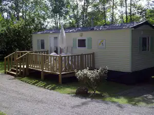 Mobilheim mit Holzveranda und Eingangstür, umgeben von Bäumen im Campingbereich, mit Kiesweg und Rasen.