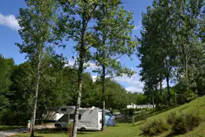 Wohnwagen und Zelt auf einem Campingplatz im Grünen, umgeben von Bäumen und einem grasigen Hang.