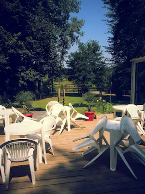 Weiße Gartenstühle auf einer Holzterrasse am Campingplatz, im Hintergrund Bäume, Rasen und ein umzäunter Bereich.