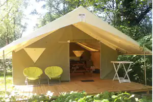 Zelt mit Holzterrasse, zwei gelben Campingstühlen und Tisch im Schatten der Bäume.