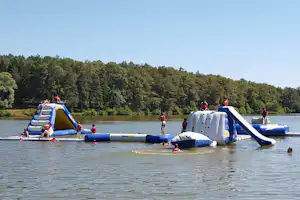 Aufblasbare Wasserbahn auf dem See, Kinder auf Luftmatten und Rutschen, im Hintergrund dichter Wald und ruhiges Wasser.