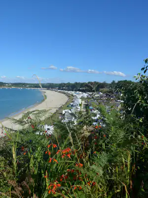 Blick vom Ufer auf einen Campingplatz am Meer mit Strand, Wohnwagen und Zelten entlang des Küstenwegs.