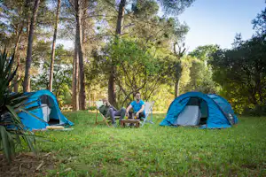 Zwei blaue Zelte auf einer Wiese im Wald, daneben Campingstühle und ein kleiner Tisch mit Getränken.