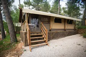 Glamping-Zelt mit Holztreppe, Seitenfenstern und Vorhang, auf Kiesplatz im Wald zwischen Bäumen.